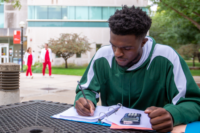 A Black male student studies over a textbook at an outdoor table on the IU South Bend campus.
